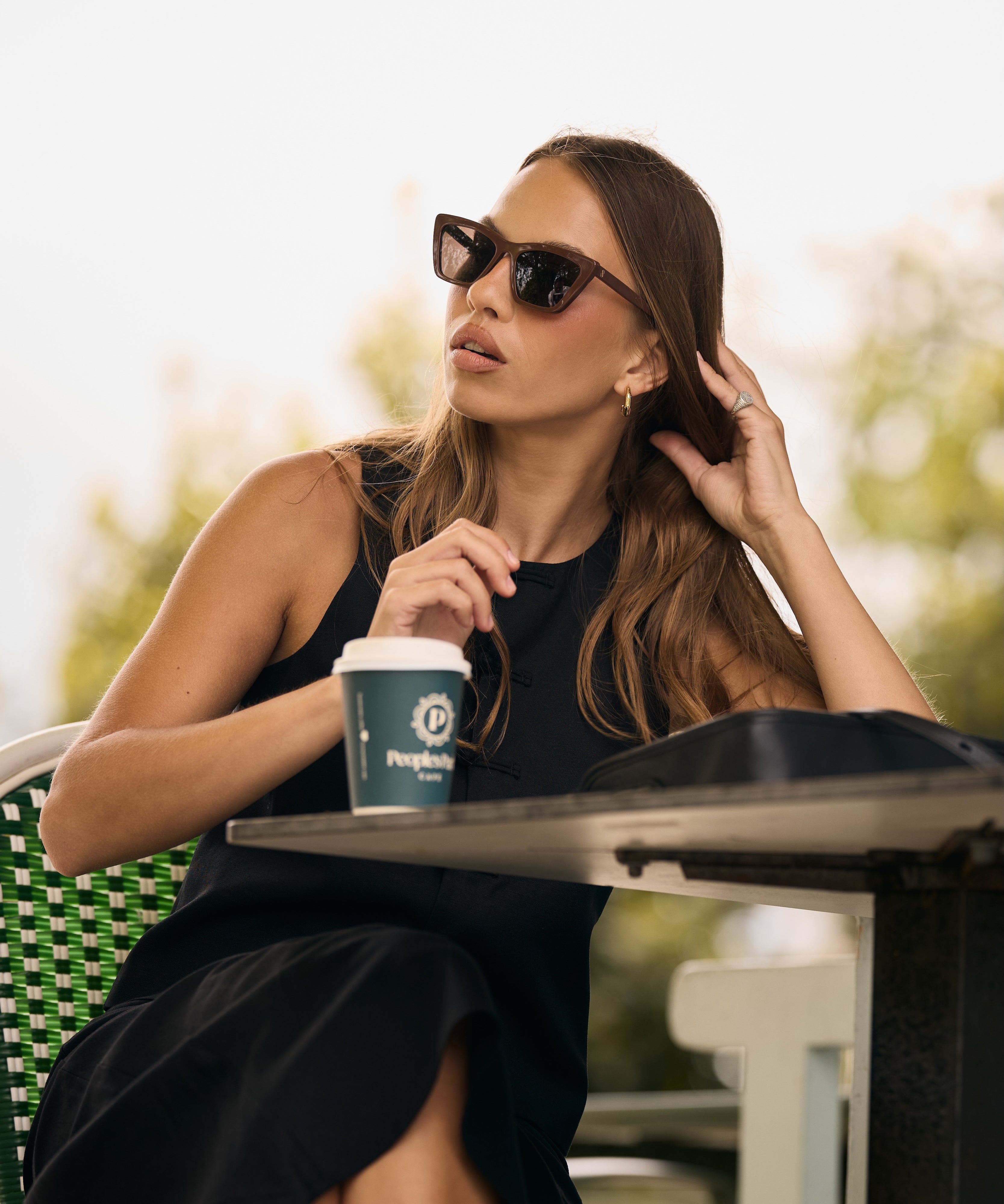 Woman sitting outdoors with a coffee cup, wearing sunglasses and a black dress.