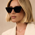 Woman wearing black sunglasses and a light-colored blazer against a neutral background