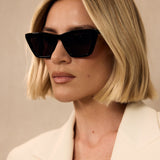Woman wearing black sunglasses and a light-colored blazer against a neutral background
