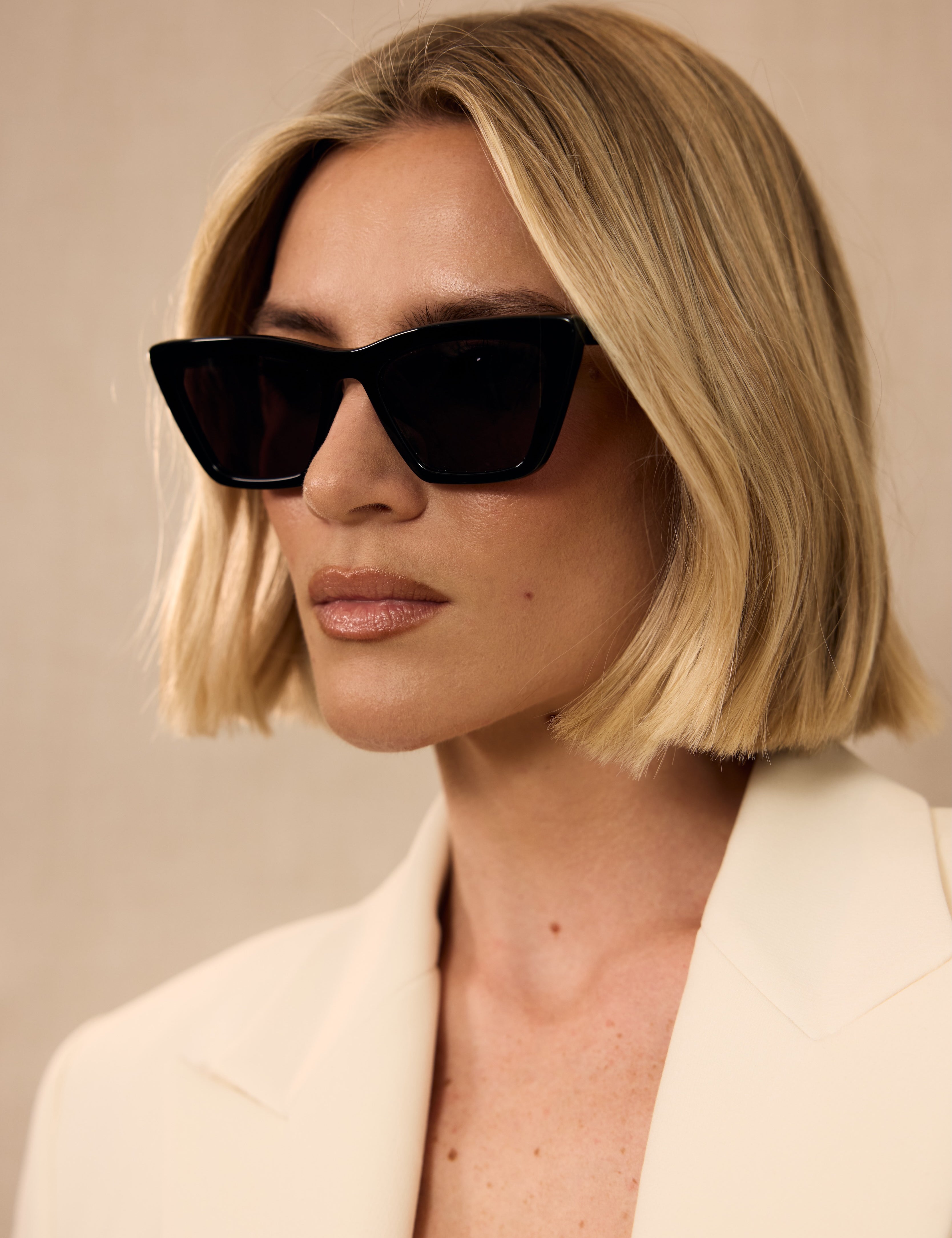 Woman wearing black sunglasses and a light-colored blazer against a neutral background