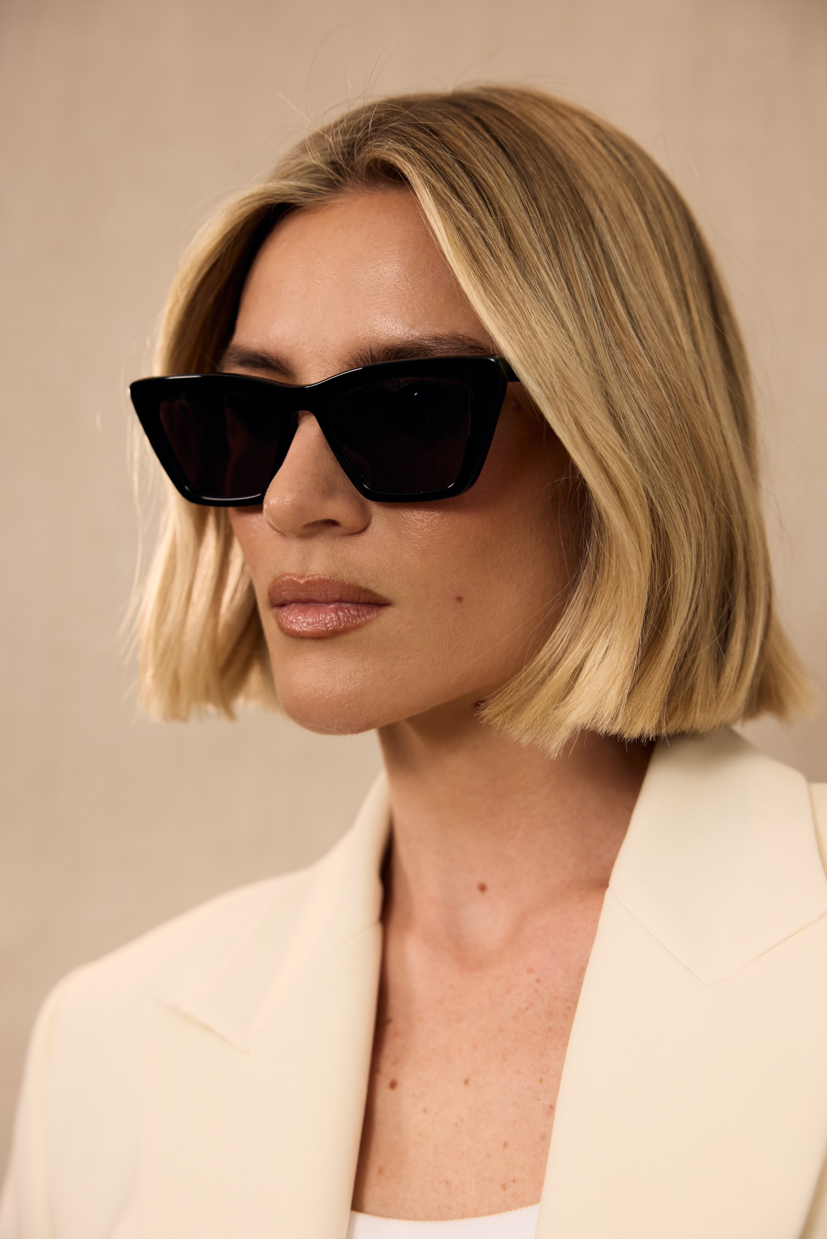 Woman wearing black sunglasses and a light-colored blazer against a neutral background