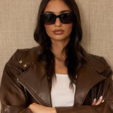 Woman wearing a brown leather jacket and sunglasses against a beige background