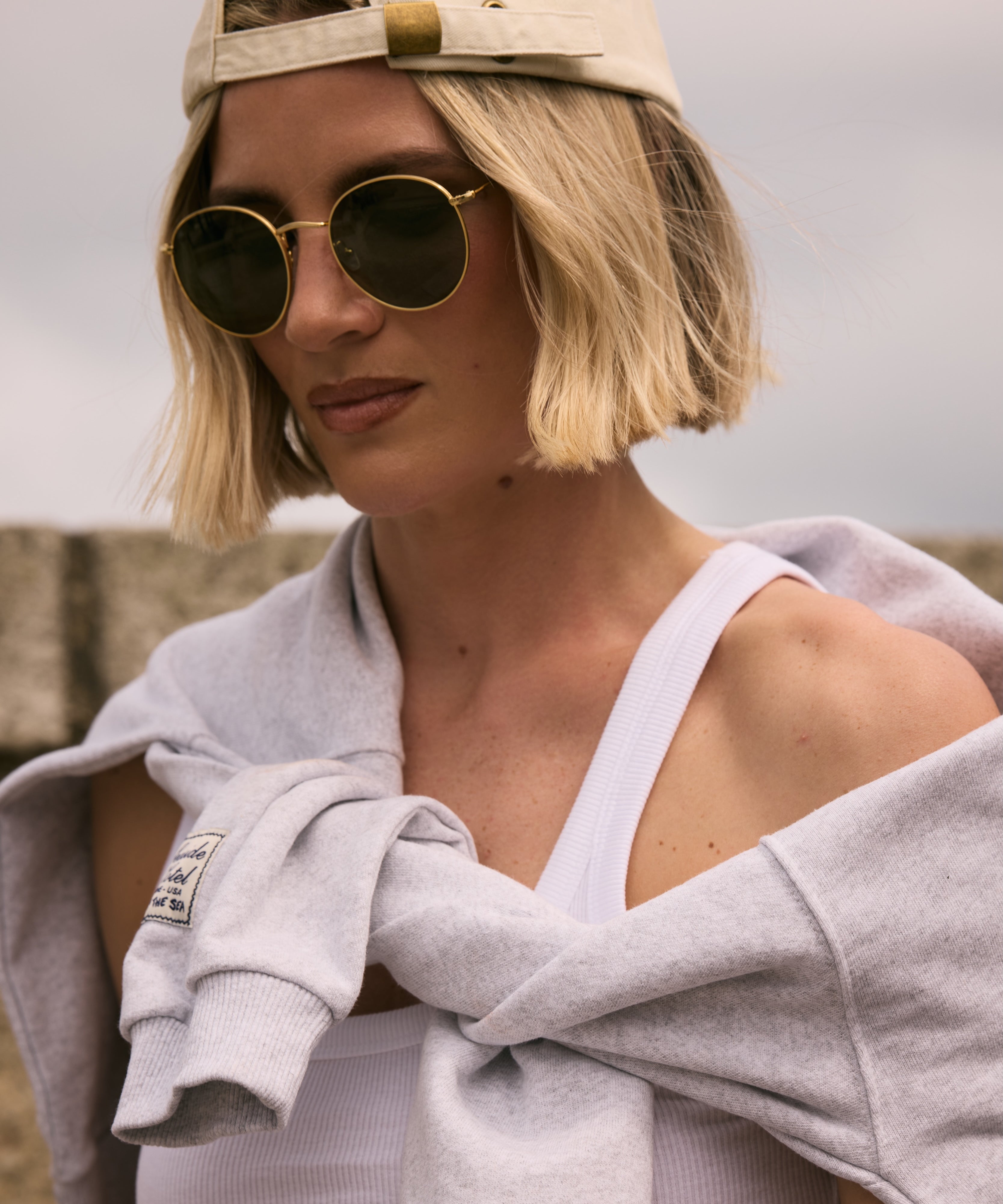 Person wearing a cap and sunglasses with a gray towel draped over their shoulder against a neutral background