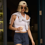 Woman in casual outfit with sunglasses and a cap, holding a drink, standing in front of a door.